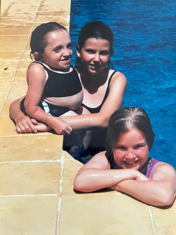 Teenage Stella sits on the edge of a pool with wet hair. Her sister Romy stands in the pool with her arms around Stella. Her other sister Madi smiles at the camera from in the pool.