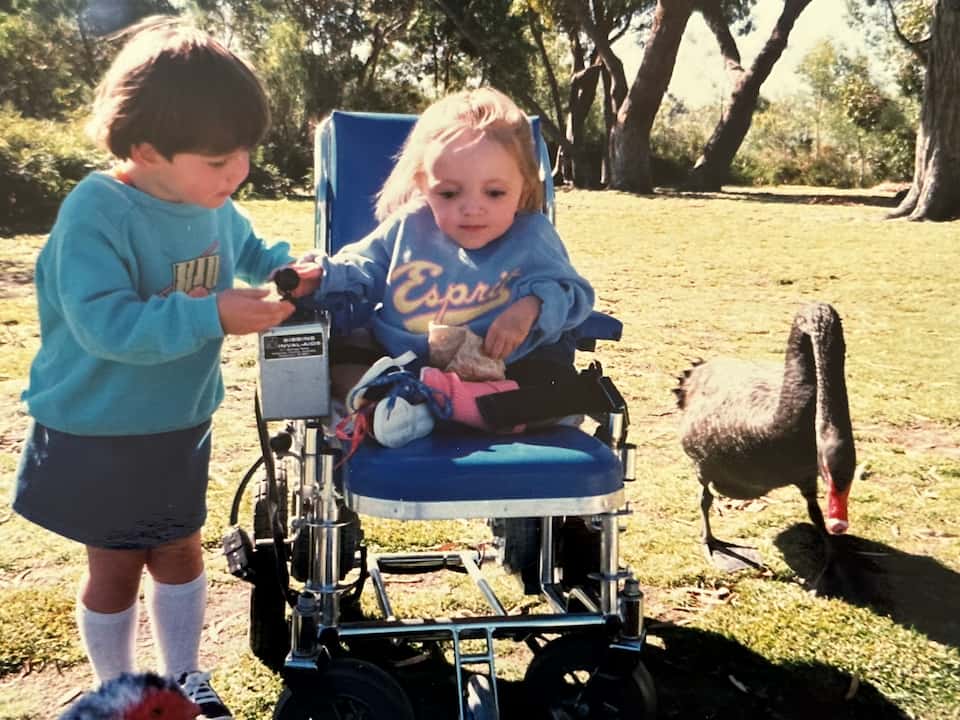 4 year old Stella sits in her electric wheelchair while her sister touches the chair's joystick. They are in a park next to a black swan pecking at the ground.