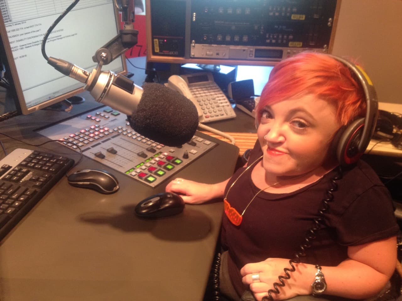 Stella is in a radio studio with a large microphone in front of her. She's behind the control panel and wearing headphones.