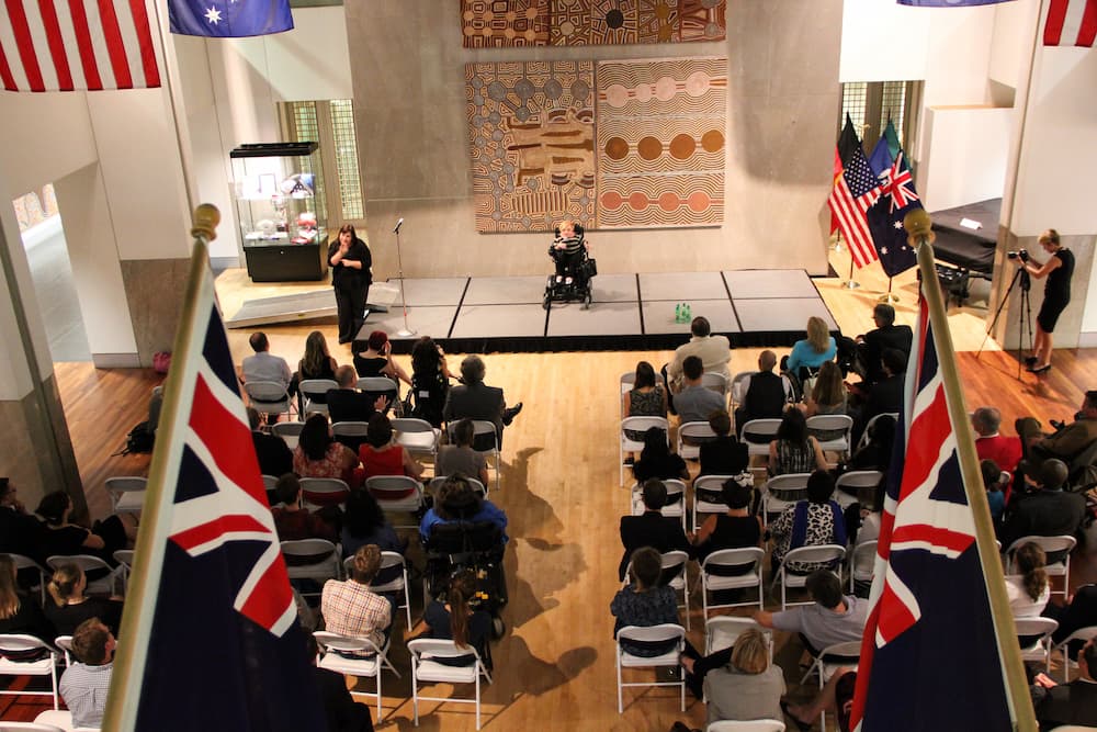 The camera looks down at Stella on a stage addressing the room. Australian flags are in the foreground and a sign language interpreter is to her right. An American flag is also visible. This is at the Australian Embassy in Washington DC.