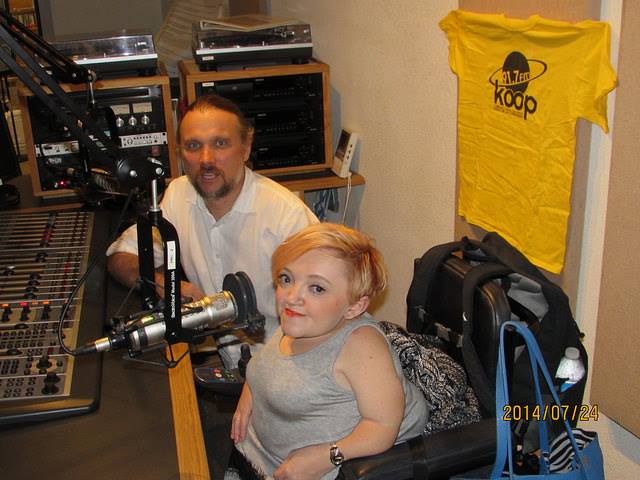 Stella is in a radio studio with a man sitting beside her. A prominent T Shirt behind her on the wall says "91.7 Koop"