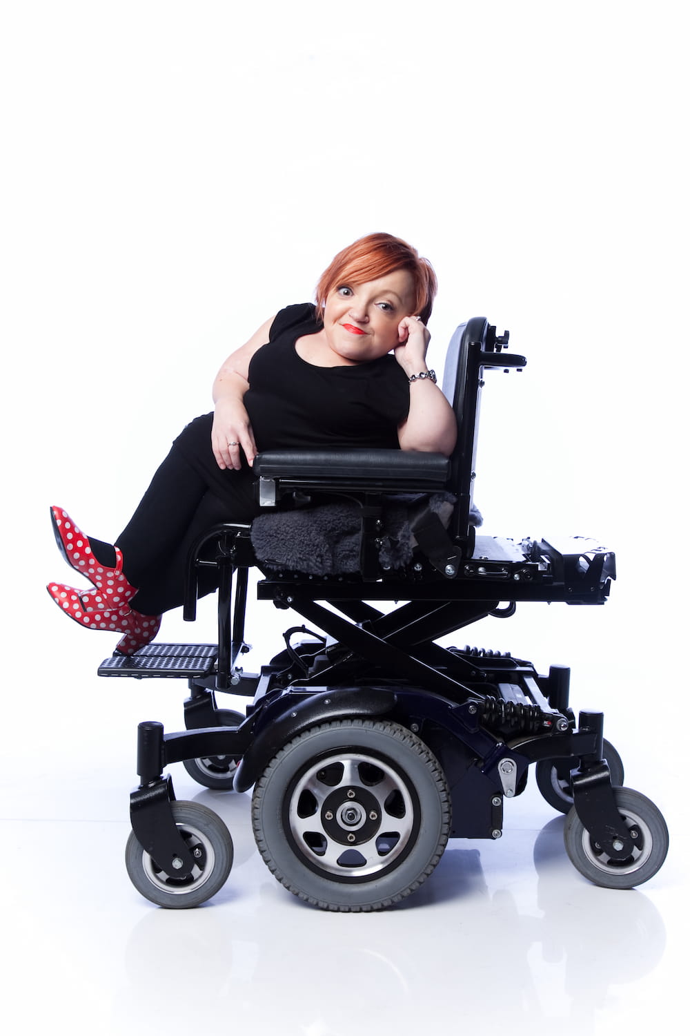 Studio photo of Stella relaxing in her wheelchair smiling at the camera. She's wearing all black except for red shoes with white polka dots.