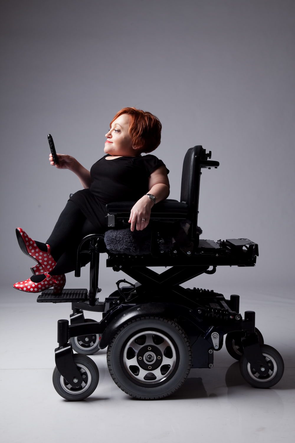 Studio photo of Stella in profile looking at her phone. She's wearing all black except for red shoes with white polka dots.