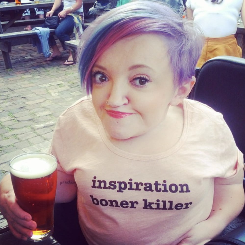 Stella has purple hair and wears a shirt that says "inspiration boner killer". She's holding a full glass of beer.