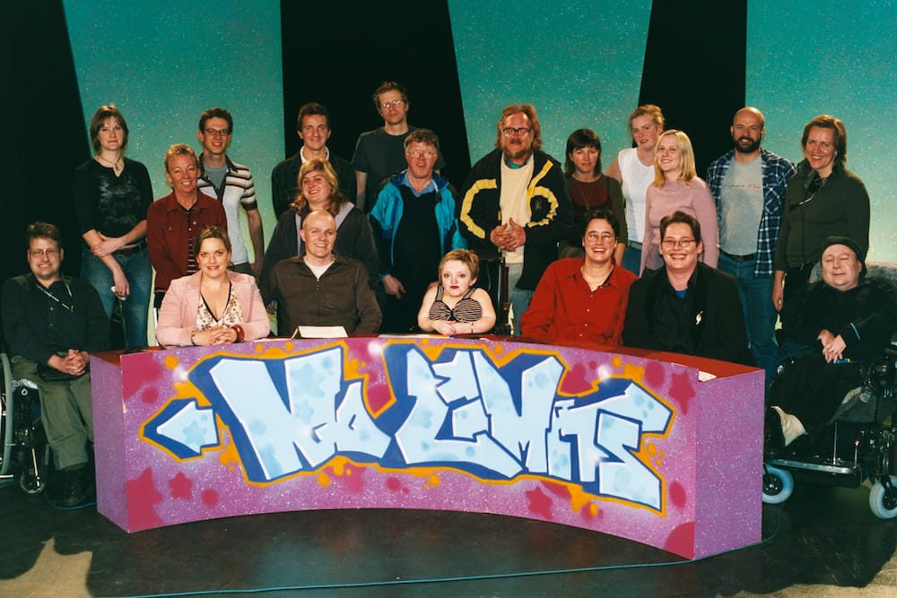 The cast and crew of No Limits - 20 people sit behind a desk with No Limits written on it in graffiti style lettering. They look happy.