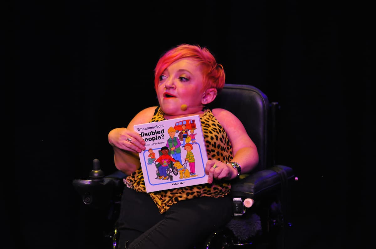 Stella holds a book titled "Who Cares About Disabled People?" She is on stage wearing a leopard print top.
