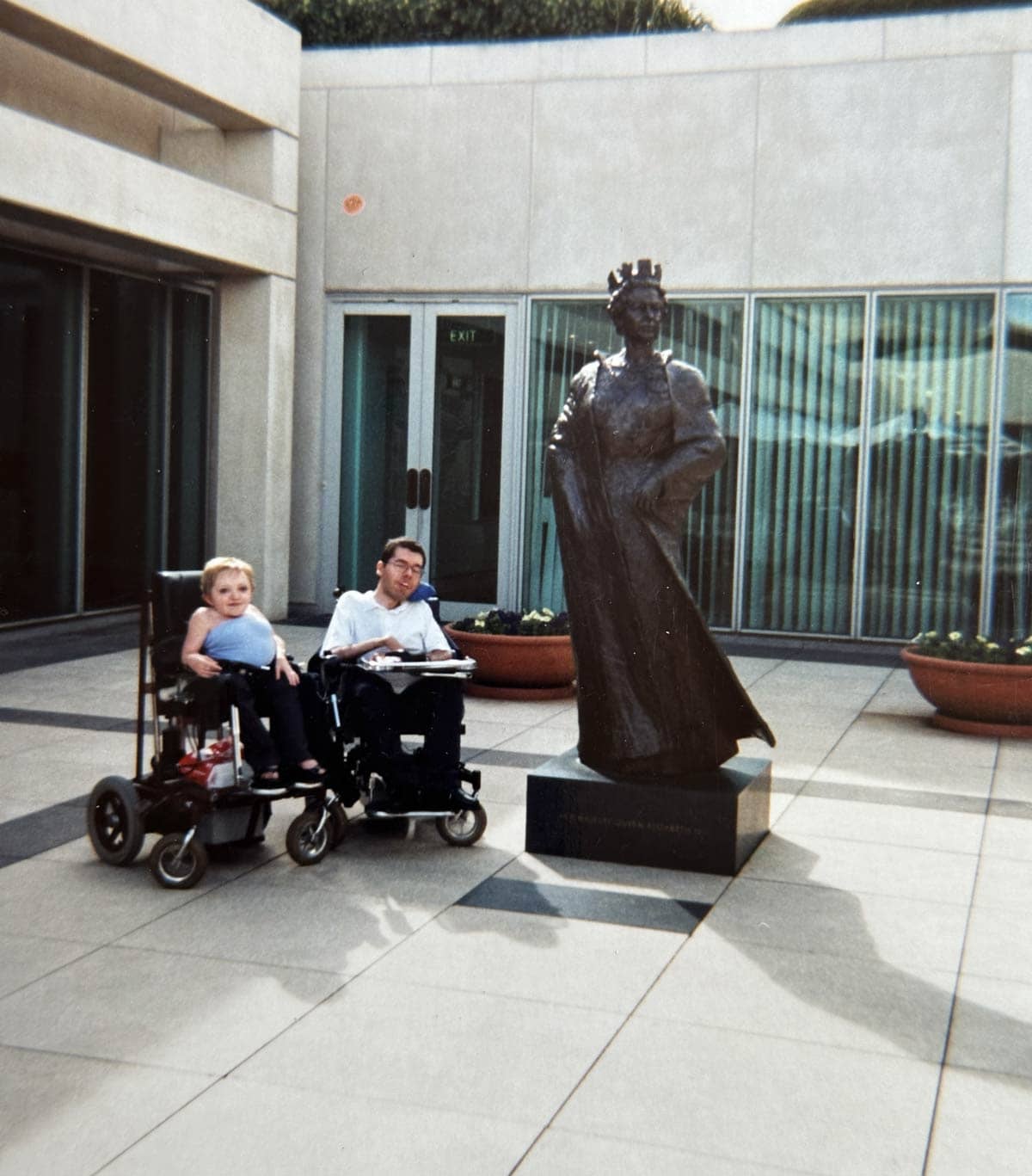 Stella and George both in electric wheelchairs pose next to a full height bronze statue of the Queen in a courtyard.