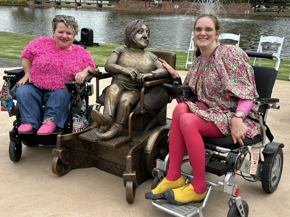 Stella Young's friends Caroline Bowditch and Stella Barton sit either side of the bronze Stella Young statue all in their wheelchairs, smiling.