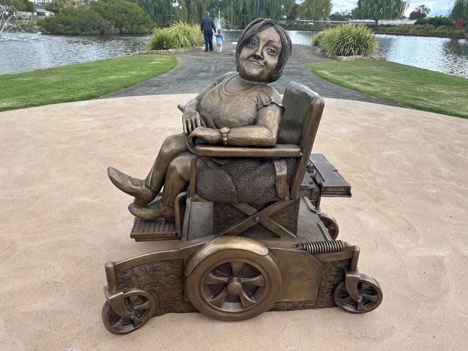 A bronze statue of Stella Young in her wheelchair sits in a park by a lake.