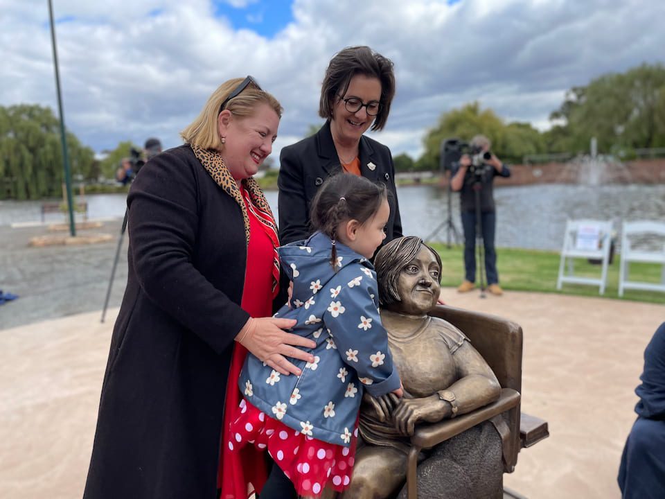 A small child climbs on the statue of Stella Young while the Minister Natalie Hutchins and an assistant look on. the minister has her hand on the child's back.
