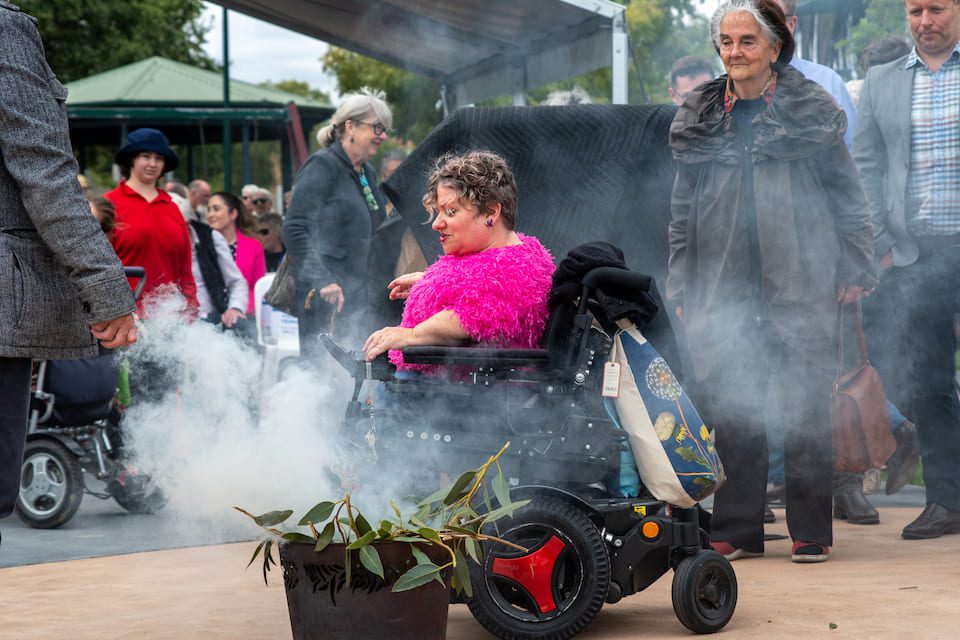 One of Stella's childhood friends wearing a pink top in a wheelchair behind the smoke from the smoking ceremony